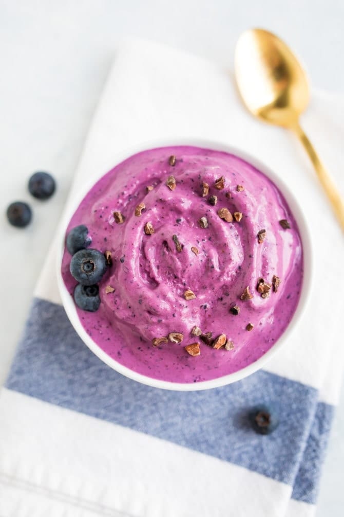 Bowl of blueberry and almond butter frozen yogurt toped with blueberries and cocao nibs. Gold spoon on the table.