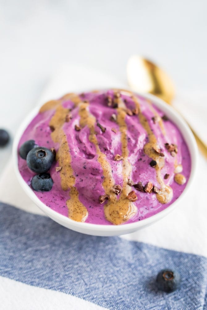 Bowl of blueberry and almond butter frozen yogurt with an almond butter drizzle, toped with blueberries and cocao nibs. Gold spoon on the table.