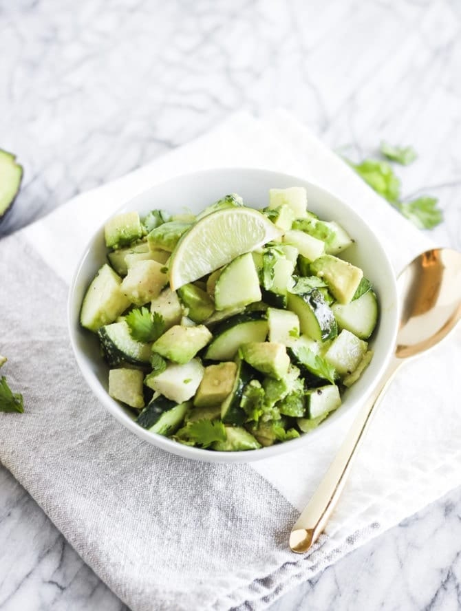 Jicama, avocado, and cucumber salad topped with a lime wedge in a bowl next to a gold spoon and on top of a folded cloth, and on top of a marble counter.