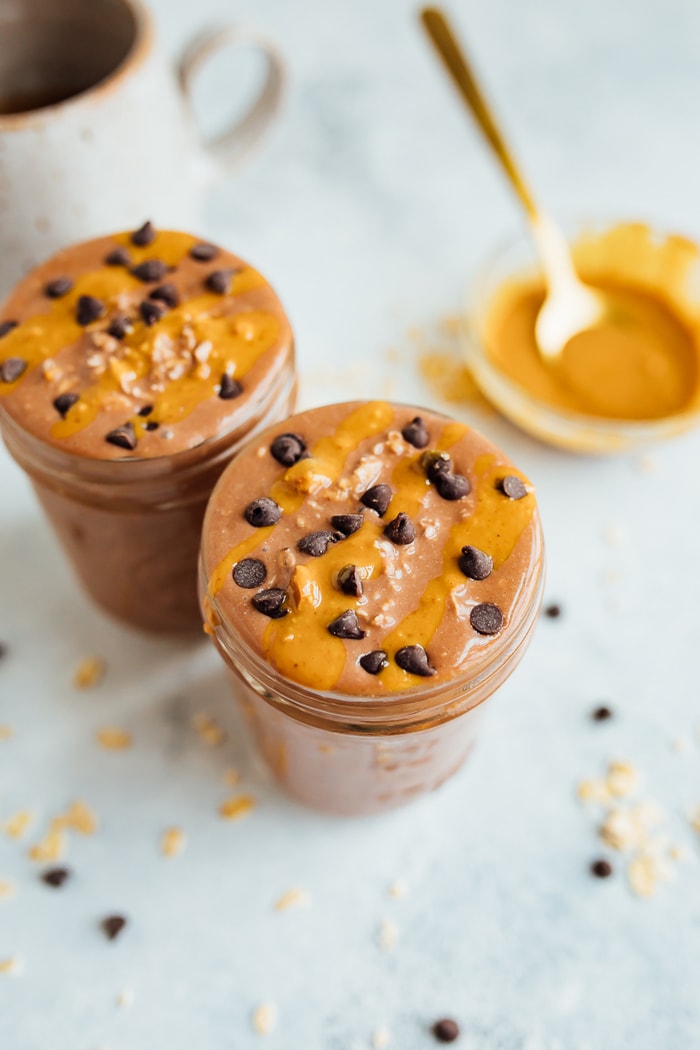 Two glass jars filled with chocolate peanut butter cup overnight oats with chocolate chips and peanut butter on top. Gold spoon in a bowl of peanut butter.