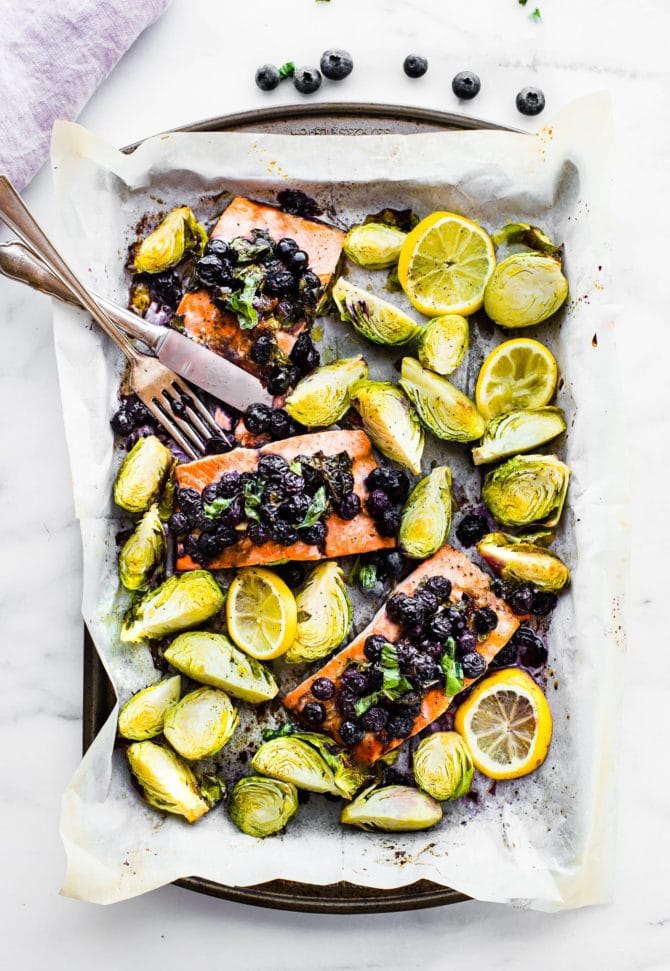 Sheet pan with parchment paper and silverware. Salmon baked with blueberries on top, Brussels sprouts and lemon.