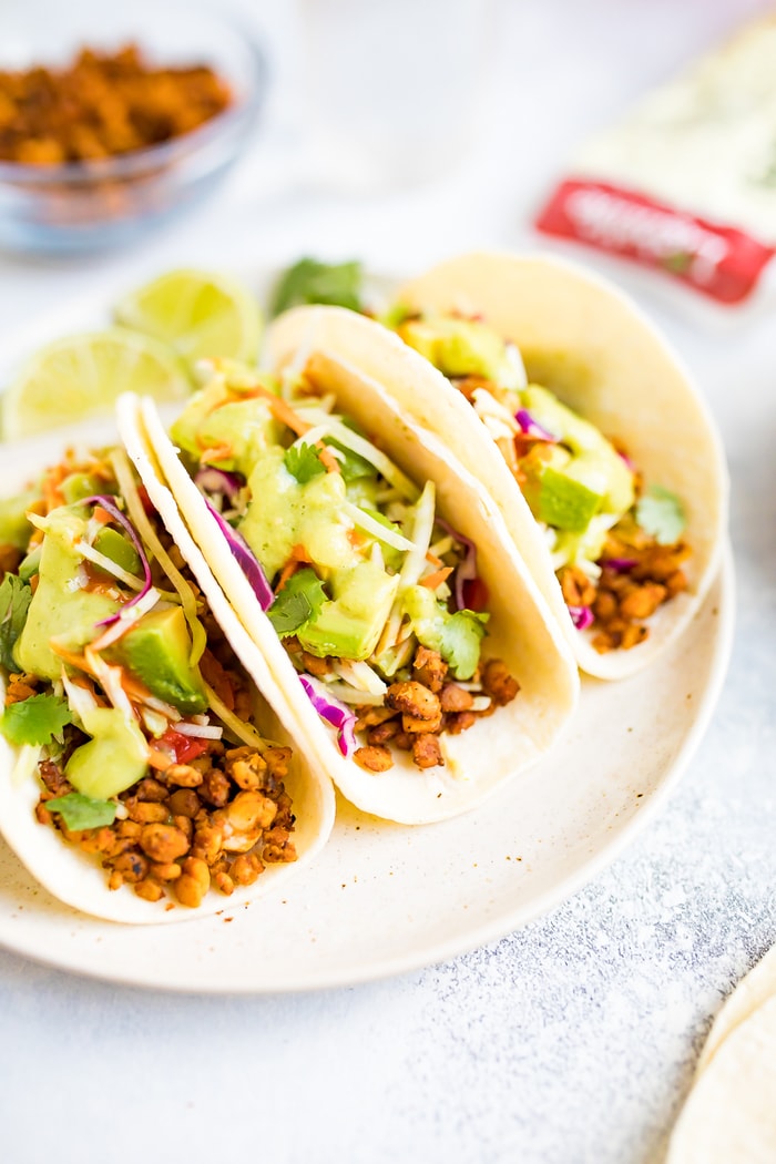 Three tempeh tacos on a white plate, topped with avocado and cilantro.