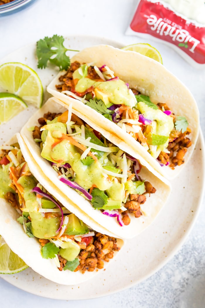 Three tempeh tacos with cilantro, avocado and tomatillo dressing on a white plate.