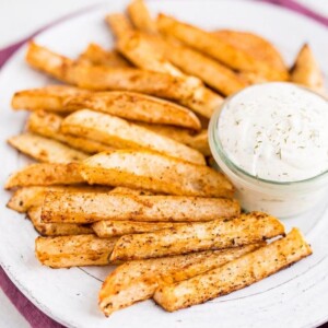 A white plate of turnip fries and a jar of ranch dip on top of a purple napkin.