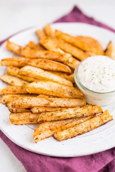 A white plate of turnip fries and a jar of ranch dip on top of a purple napkin.