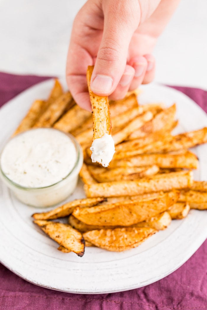 White plate of turnip fries over a purple napkin. A hand holds a fry that has been dipped in ranch dip.