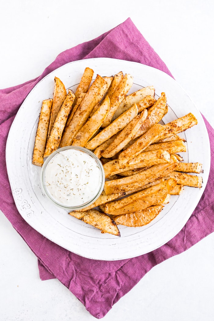 A white plate of turnip fries and a jar of ranch dip on top of a purple napkin.