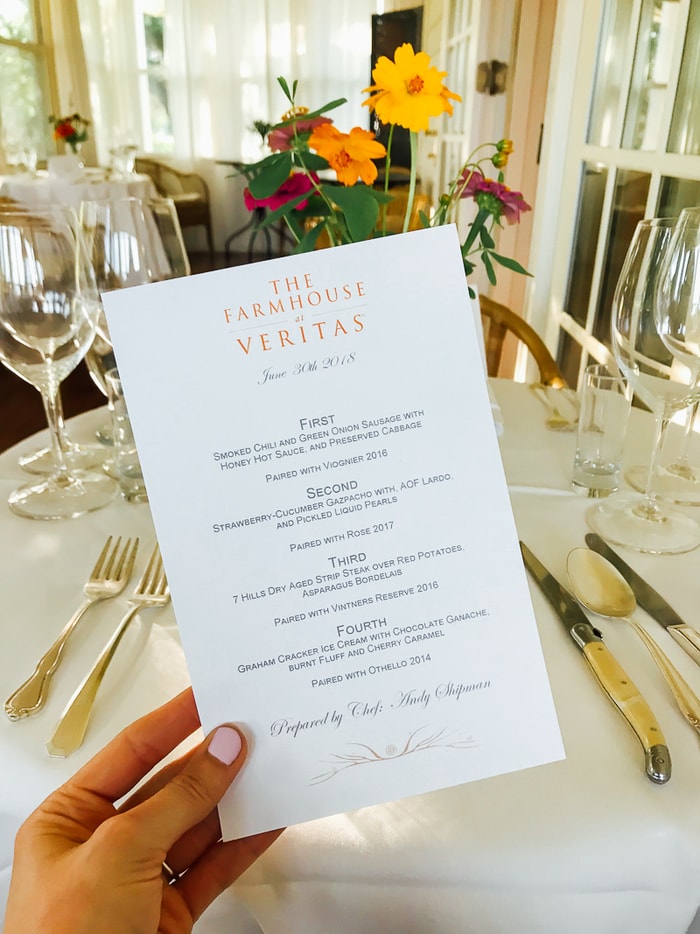 Dinner menu from The Farmhouse at Veritas with four courses. Hand holding menu with table set with silverware and flowers in the background.