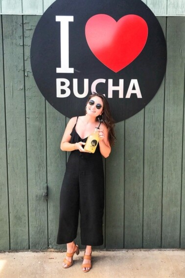 Woman holding kombucha, standing in front of a sign reading I heart bucha.