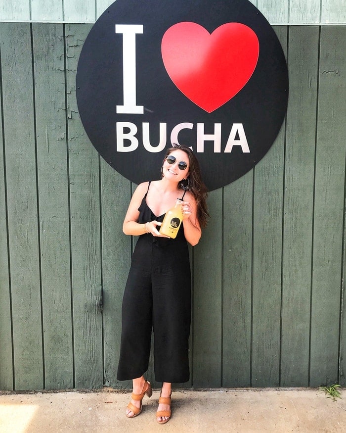 Woman holding kombucha, standing in front of a sign reading I heart bucha.