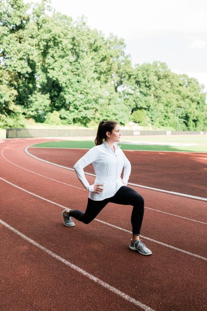 Woman doing lunges on a track, wearing leggings and a zip up pullover.