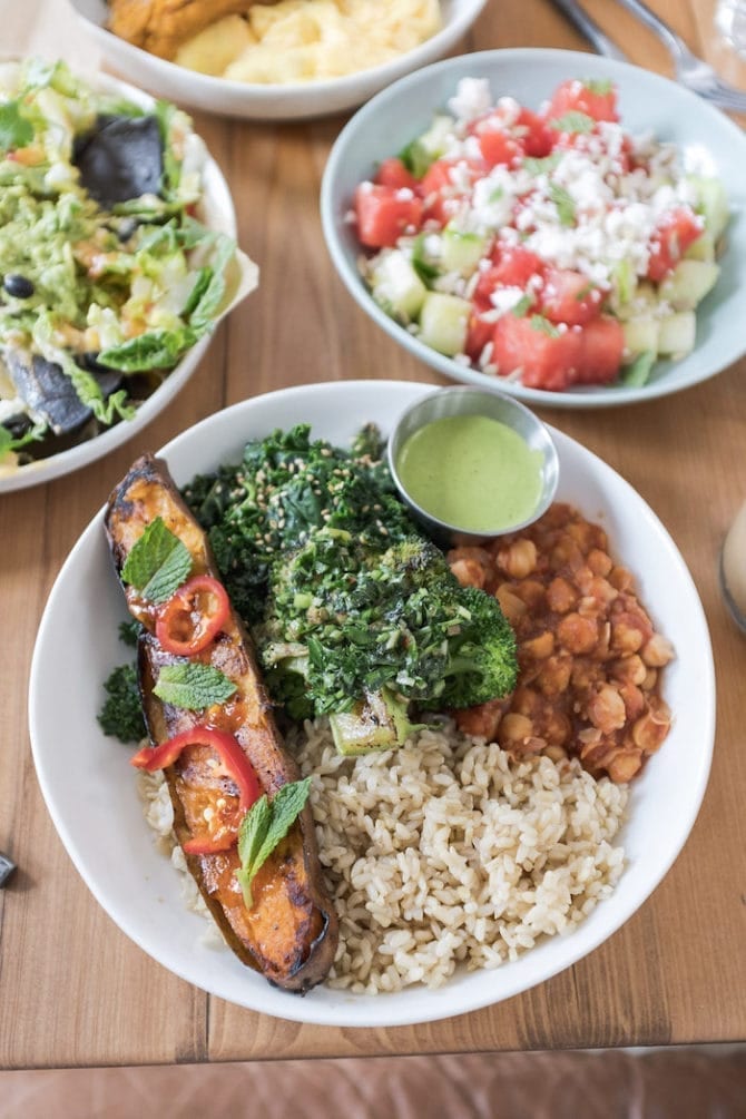 Bowl filled with sweet potato, broccoli, brown rice and chickpeas. There are other bowls of salads and guacamole in the background.