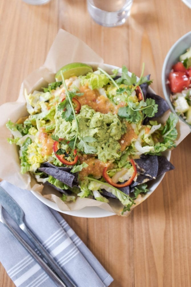 Bird's eye view of a bowl, lined with parchment paper, and filled with blue corn chips and piled with guacamole and salsa.