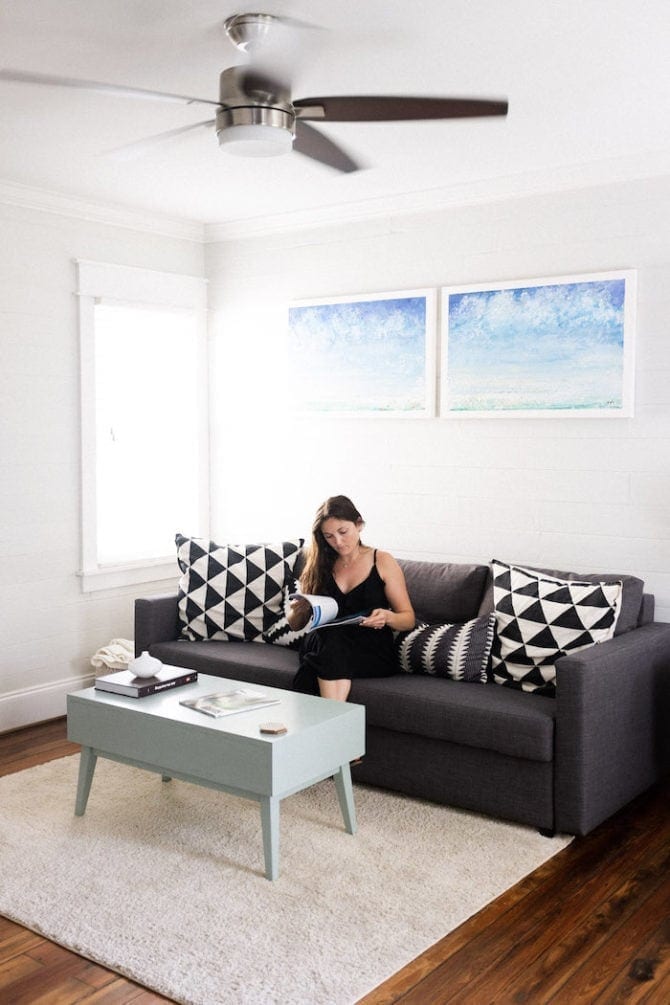 Woman sitting on a couch looking as a magazine. There is a coffee table, a rug and a couple paintings in the room. A window is beside her. The room is very clean and minimalist.