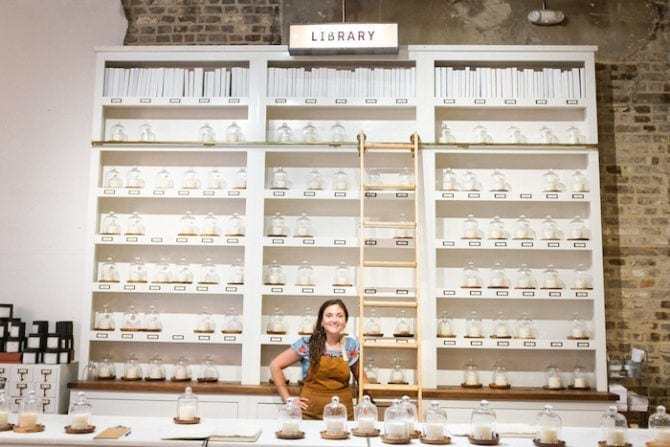 Woman wearing an apron and standing in front of a wall filled with shelves of candle waxes. A sign says "library".