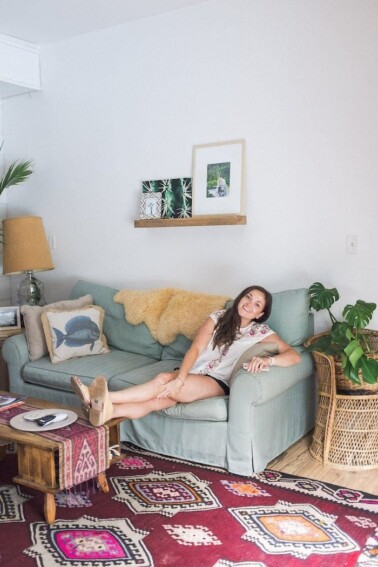 Woman lounging on a soft couch with her feet propped on a coffee table. There are woven rugs, house plants, and beach themed decor.