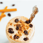 Glass jar of cinnamon raisin overnight oats, topped with cinnamon and raisins, and a silver spoon in the jar. Cinnamon sticks and raisins off to the side on the table. Text above reads "Cinnamon Raisin Overnight Oats".