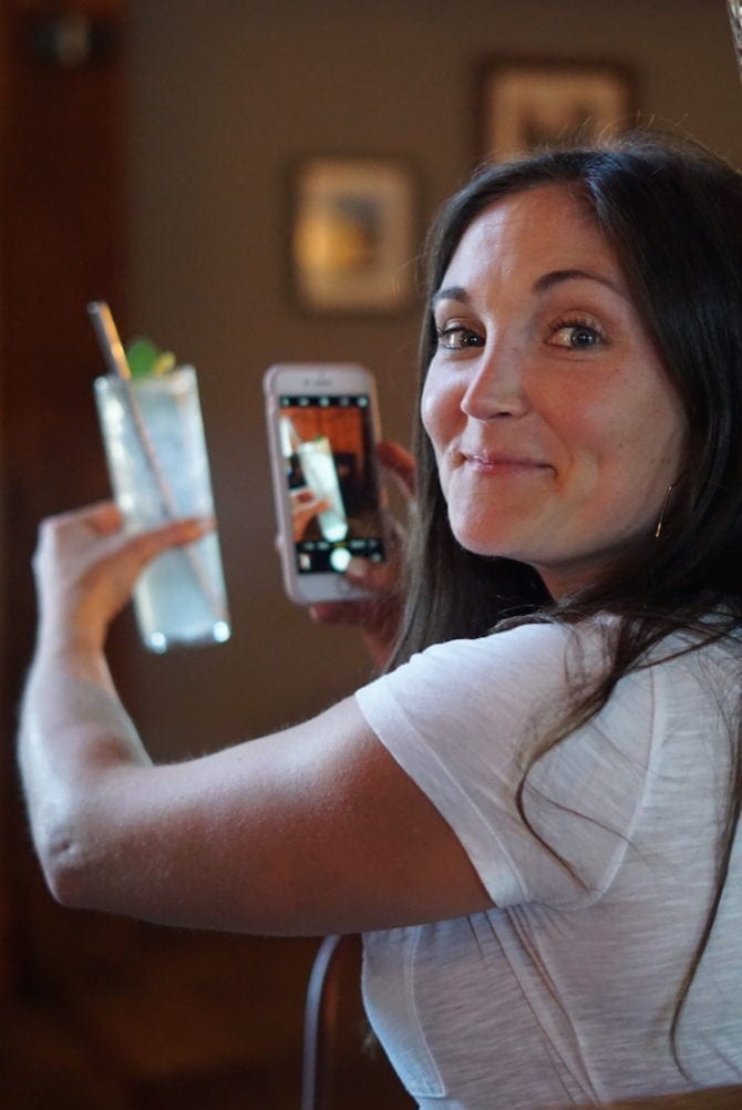 A photo of a woman taking a photo with her iPhone of her cocktail.