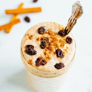 Glass jar of cinnamon raisin overnight oats, topped with cinnamon and raisins, and a silver spoon in the jar. Cinnamon sticks and raisins off to the side on the table.