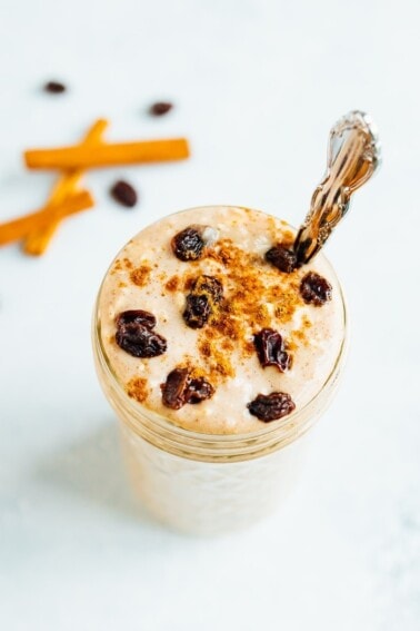 Glass jar of cinnamon raisin overnight oats, topped with cinnamon and raisins, and a silver spoon in the jar. Cinnamon sticks and raisins off to the side on the table.