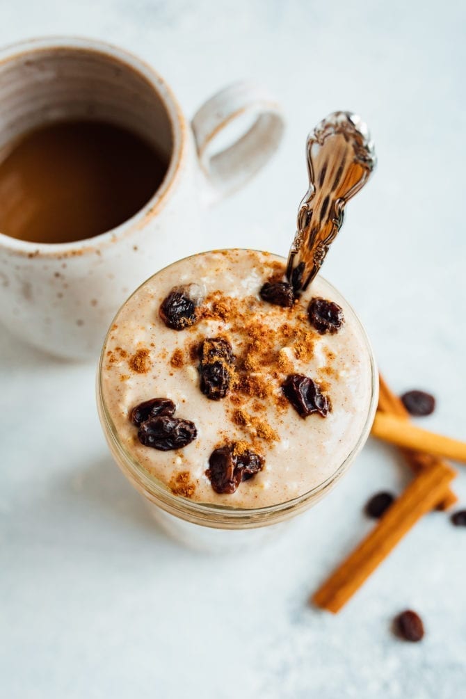 Glass jar of cinnamon raisin overnight oats, topped with cinnamon and raisins, and a silver spoon in the jar. Cinnamon sticks, raisins, and a mug of coffee off to the side on the table.