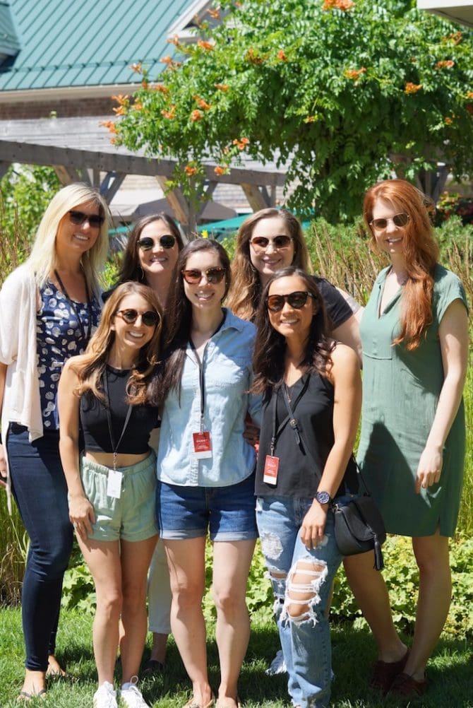 A group of 7 young women standing under a wooden roof outdoors with flowery trees behind them.