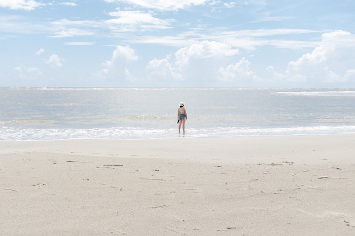 Woman in jean shorts and a hat standing in the surf of the ocean, on a beach in Charleston, SC. Sky is blue with a few light clouds.