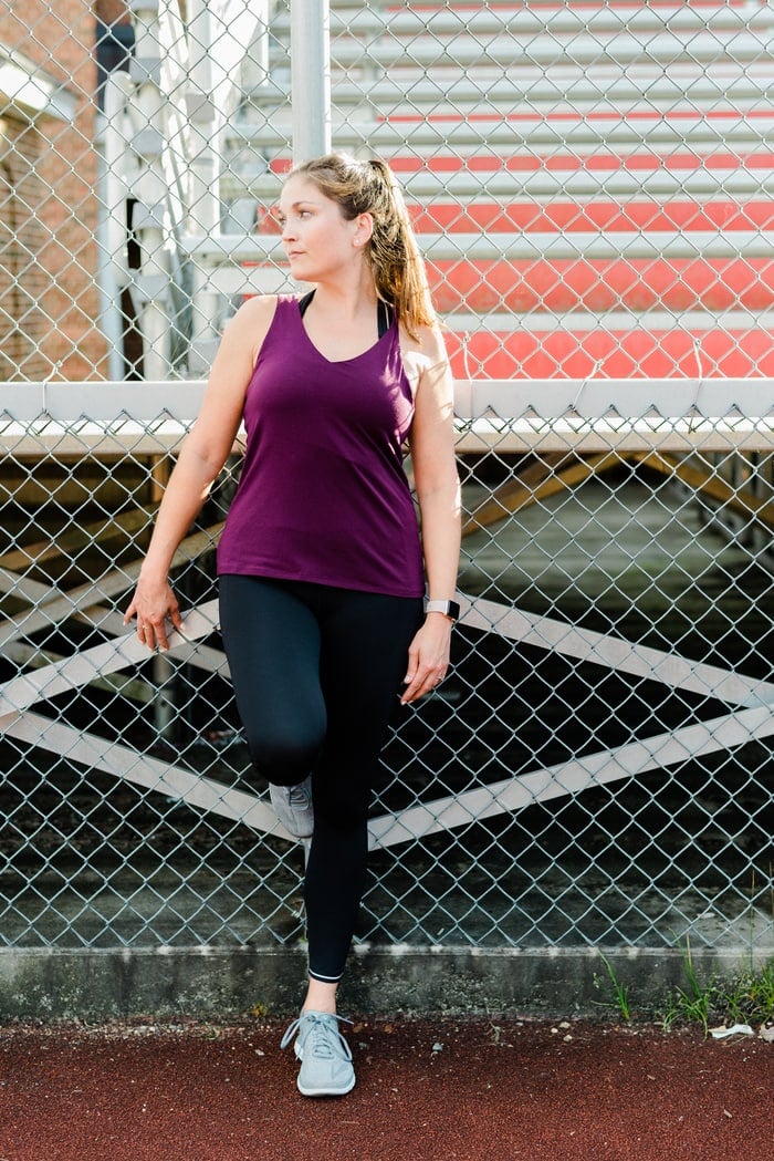 Woman standing against a chainlink finch in front of bleachers wearing Athleta running clothing.