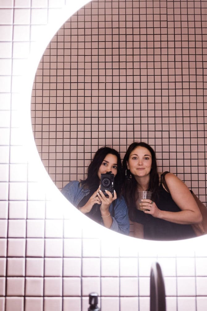 Two women taking a mirror selfie. Mirror is round and surrounded by light. The wall is pink and tiled.