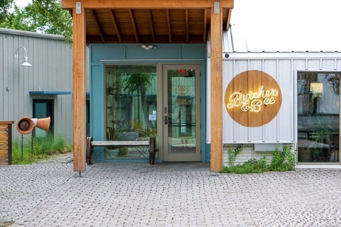 Outside of the Butcher & Bee restaurant in Charleston, SC. There are glass windows and a door, and a mixture of wood and metal on the modern building.