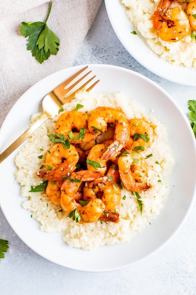 Plate of cauliflower cheese grits topped with blackened shrimp and chipped fresh parsley. A fork is resting on the plate.