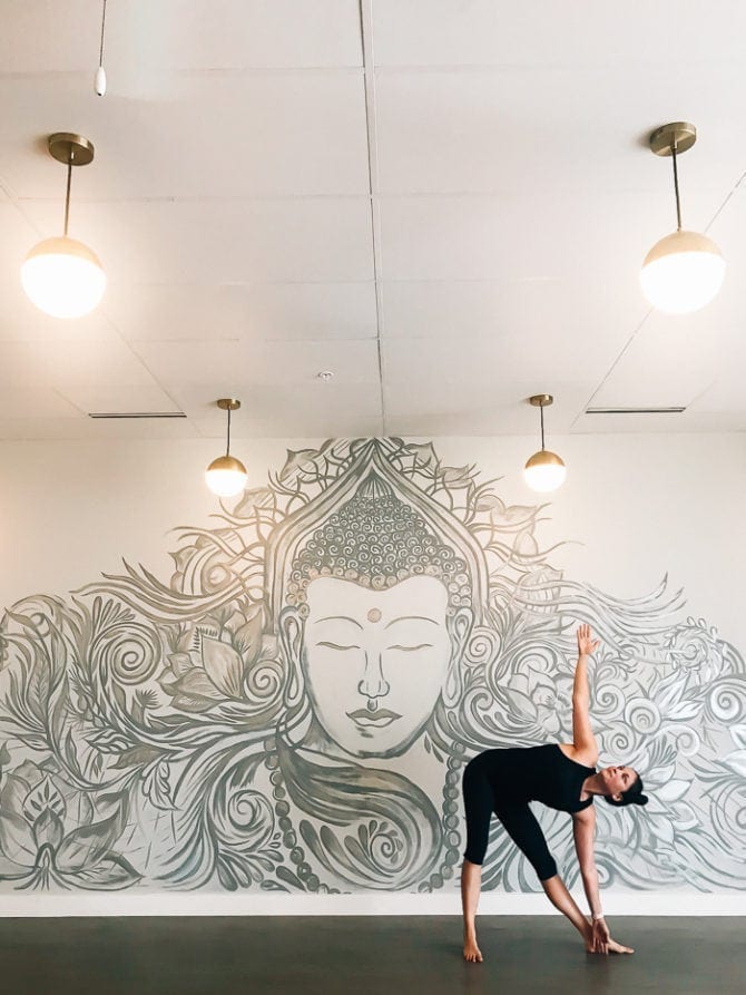A woman doing a yoga pose in front of a wall with a mural of Buddha. Round lights hang from the ceiling.