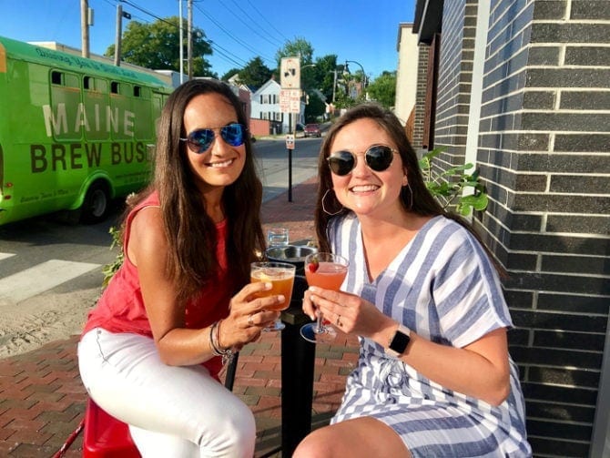 Two women in sunglasses holding cocktails at an outdoor table with the Maine Brew Bus in the background.