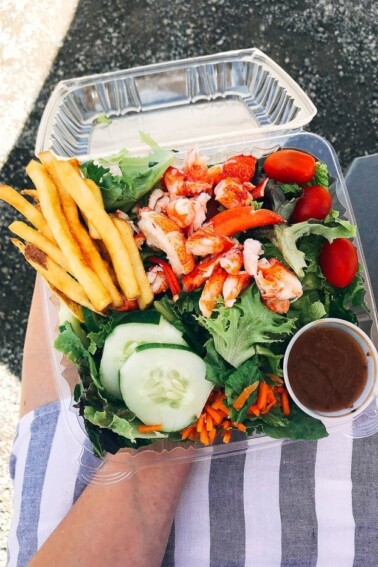 Woman holding a plastic container that is filled with salad topped with cucumber, carrots, tomatoes, lobster, salad dressing and French fries.