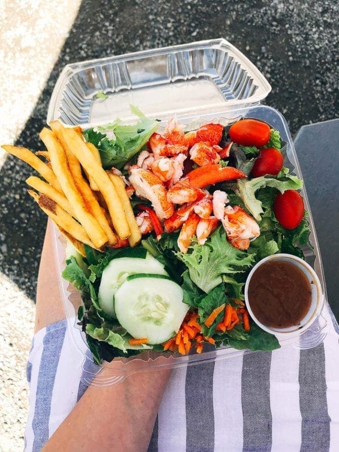 Woman holding a plastic container that is filled with salad topped with cucumber, carrots, tomatoes, lobster, salad dressing and French fries.