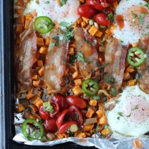 Sheet pan with baked sweet potato cubes, jalapeño, eggs, tomatoes, and bacon slices.