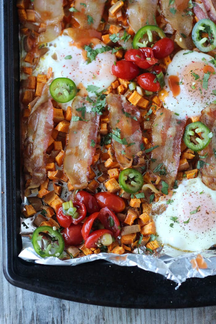 Sheet pan with baked sweet potato cubes, jalapeño, eggs, tomatoes, and bacon slices.