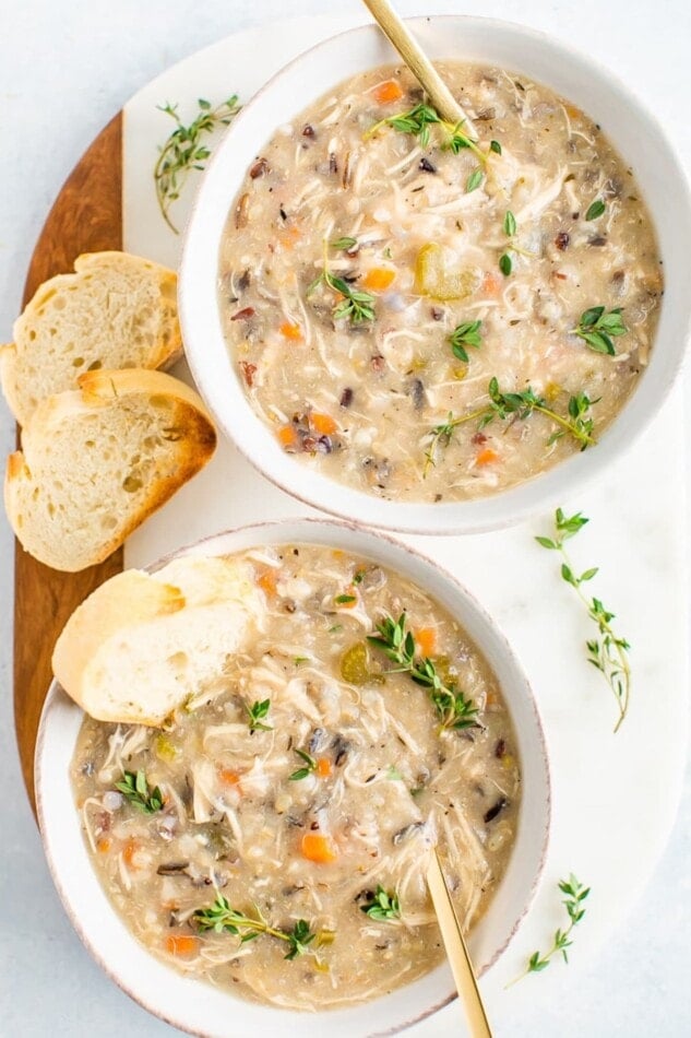 Two bowls of wild rice chicken soup served with fresh thyme and slices of bread. Two spoons and a cloth napkin are around the soup bowls.
