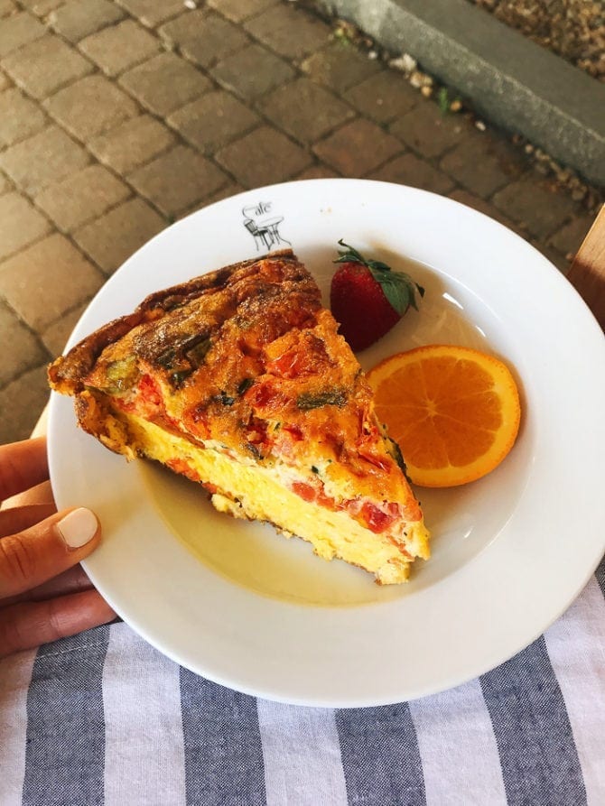 PIN THIS--> A Trip to York, Maine Woman's hand holding a plate with a slice of quiche with peppers, green veggies and cheese. A slice of orange and a strawberry are on the plate.