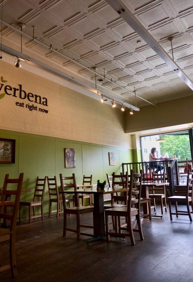 Inside of a restaurant with wooden tables and chairs, an antique metal ceiling and "verbena, eat right now" on the wall.