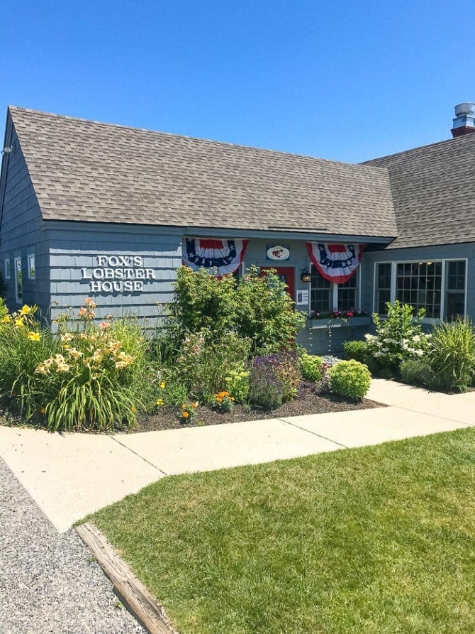 PIN THIS--> A Trip to York, Maine Photo of Fox's Lobster House, a wood shingled restaurant with a grassy lawn, garden, and American flag banners.