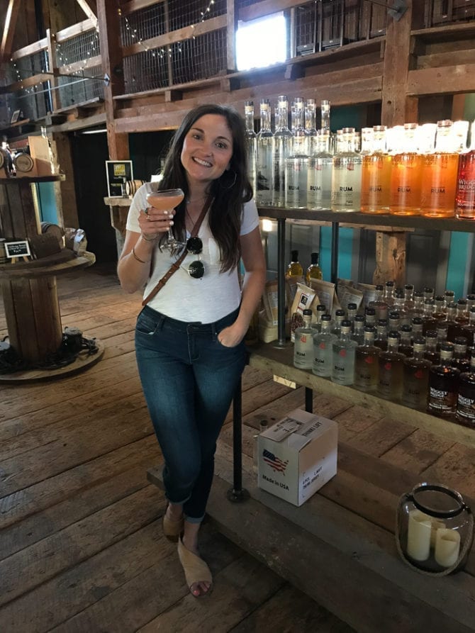 PIN THIS--> A Trip to York, Maine A woman in a t-shirt and jeans posting with her cocktail in front of liquor shelves in a distillery. The floors and walls are wooden.