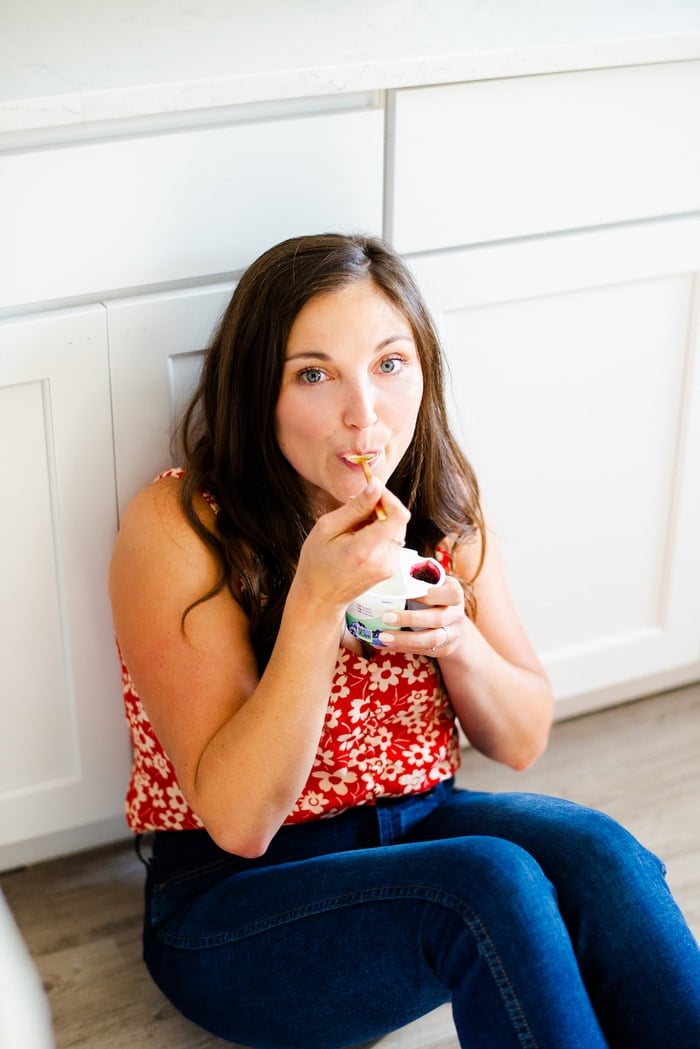 Woman sitting on the floor of a kitchen eating a cup of yogurt.