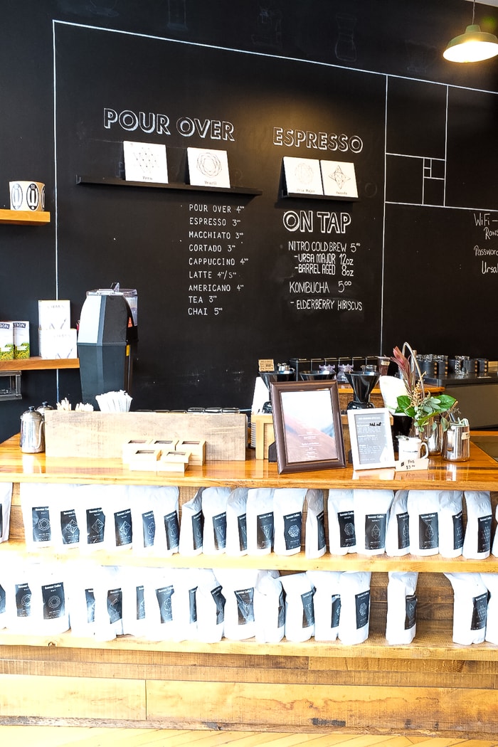 Wood shelves of coffee bags in front of a chalk board coffee shop wall menu.