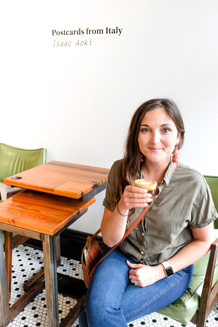 Woman sitting in a chair next to a wood side table, holding coffee. The wall has the words "Postcards from Italy/Isaac Aoki" behind her.