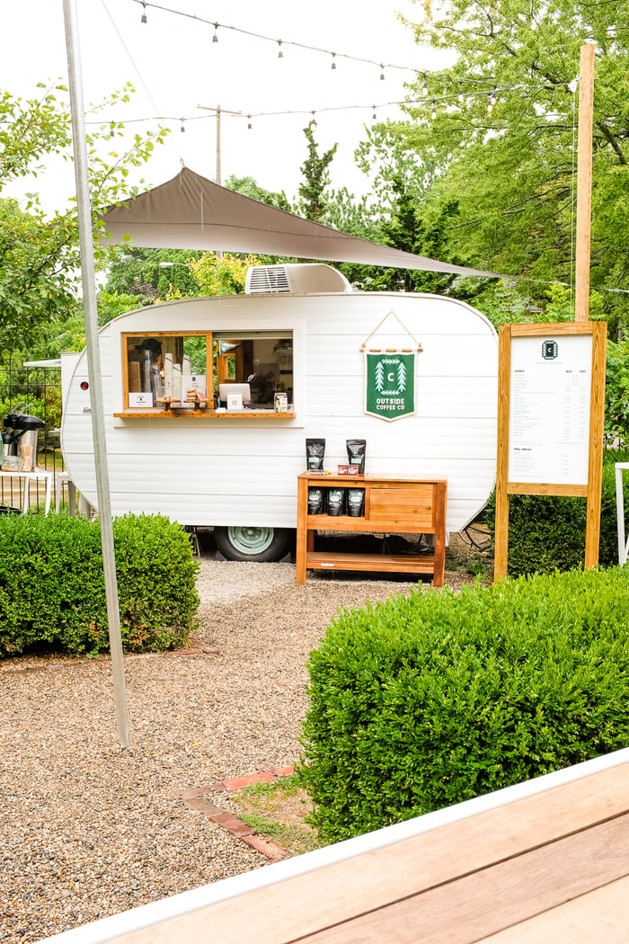 Airstream trailer that is a coffee shop trailer with a menu and string lights above. Outside in a lush courtyard.