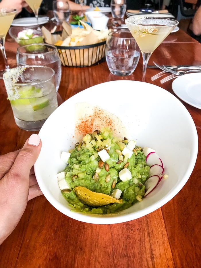 Woman holding a bowl of guacamole at a restaurant table. The guacamole is topped with seeds, jicama and radish slices.