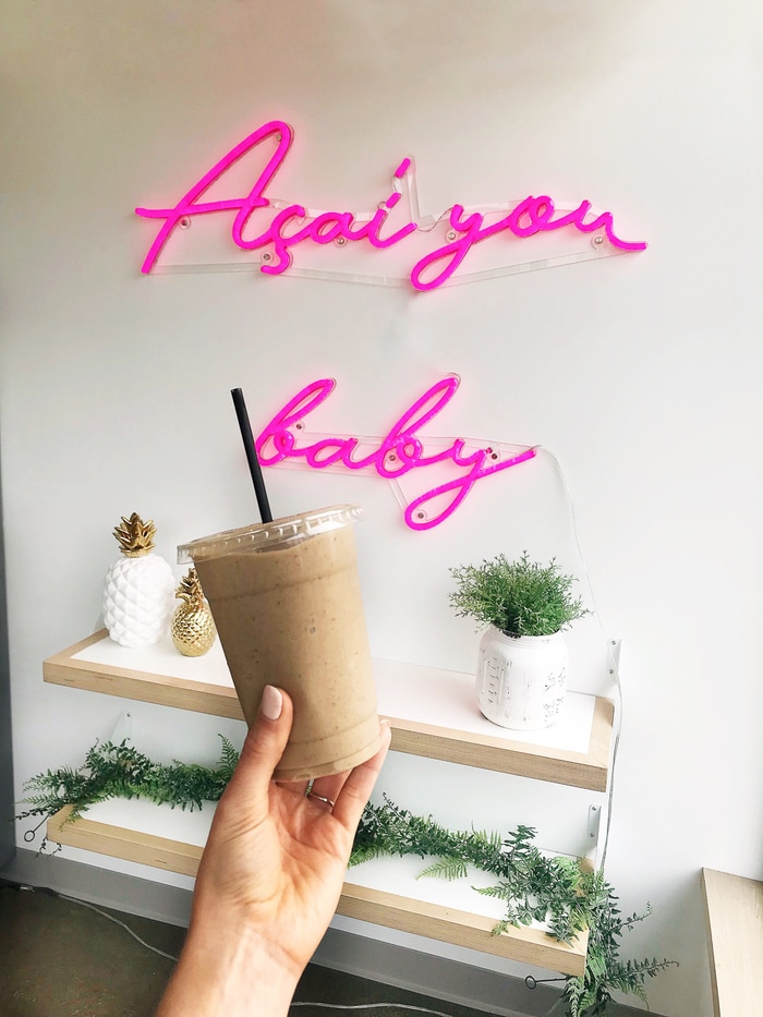 Woman's hand holding a protein smoothie in front of a wall that has a neon sign that says "Acai you baby" and some plants on wall shelves.