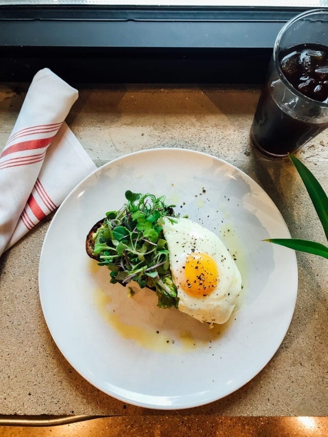 plate of avocado toast topped with micro greens and a fried egg. A napkin and glass of iced coffee are next to the plate.