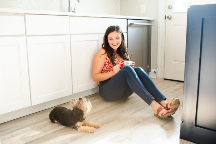 Woman sitting on the floor of her kitchen eating yogurt and laughing while looking at her small dog next to her.